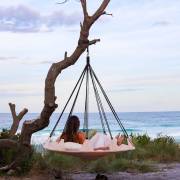 Beige open hangout hung from a tree strap on a branch at the beach, with a woman lying back looking at the sea
