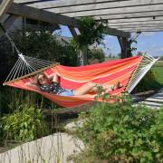 Woman relaxing in the red and orange spreader bar hammock under a pergola