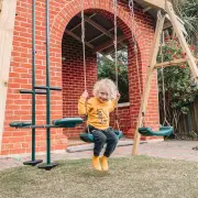 Child on a swing seat between the glider and the second seat, in front of a brick house