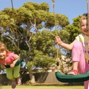 One girl lying over a green swing seat and another standing on one, holding the ropes