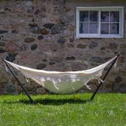 Beige fringe hammock on its steel stand on a lawn beside a stone wall