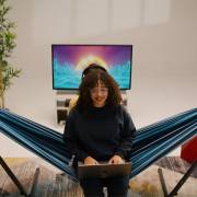 Woman sitting in the mixed blues hammock on its steel stand indoors with a laptop