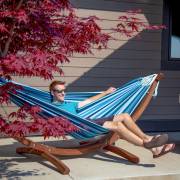 Man lying in the mixed blues hammock on its wood arc stand beside a maple tree