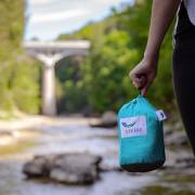 Turquoise parachute hammock packed into its stuff sack, held by a hand at a creek