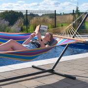 Woman reading in a multi colour spreader bar hammock on the three beam stand beside a pool